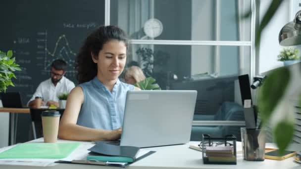 Glada unga kvinna som använder laptop skriva medan kollegor arbetar i bakgrunden i delat kontor — Stockvideo