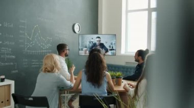 Çalışanlar dijital ekrana bakıp video çekiyor erkek meslektaşlarını dinliyor ve ofiste el sallıyorlar.