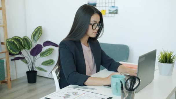 사무실에서 노트북으로 일하는 아시아인 여자 직원 이 커피를 마시며 서류를 보고 있습니다. — 비디오