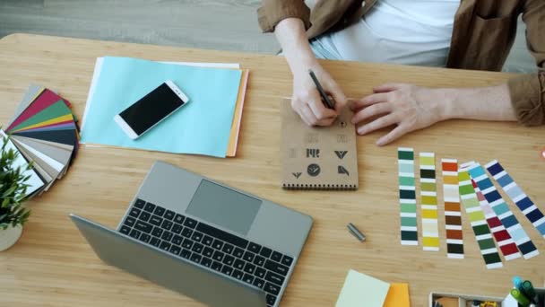 Nagy látószögű kilátás férfi kéz tervező vázlatos logó szimbólumok és gépelés laptop dolgozik az irodában — Stock videók