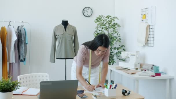 Pretty Asian girl fashion designer is drawing fashionable clothes on paper creating new collection of garments focused on creative activity in studio — Stock Video