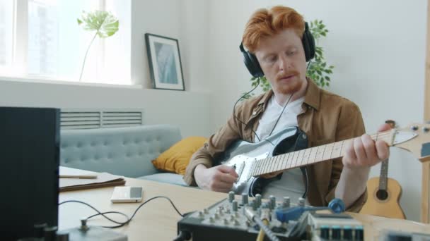Retrato de câmera lenta de músico cara atraente tocando guitarra usando fones de ouvido ajustando equipamentos em casa — Vídeo de Stock