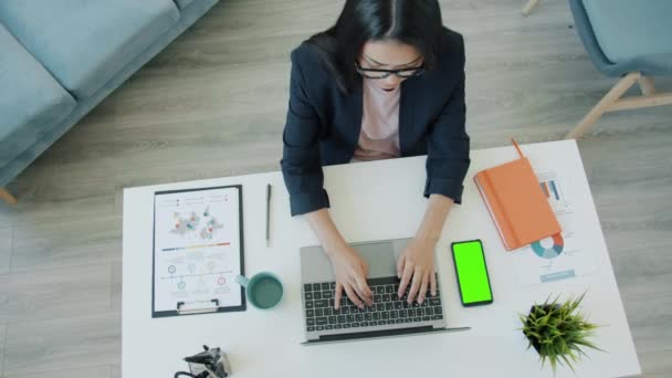 Hög vinkel syn på unga affärskvinna som arbetar hemma med hjälp av datorn och titta på chroma nyckel grön skärm smartphone — Stockvideo