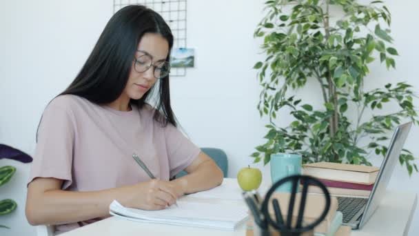 Giovane signora asiatica guardando lo schermo del computer portatile e scrivendo facendo i compiti in casa in appartamento — Video Stock