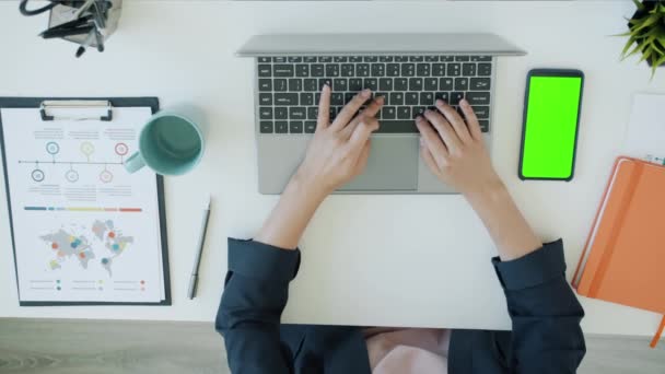 Vista dall'alto delle mani femminili che lavorano con il computer portatile in ufficio mentre lo smartphone con chiave cromatica verde è visibile sulla scrivania — Video Stock
