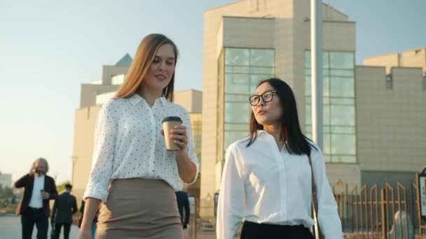 Młode biznesmenki Azjatki i Kaukaskie spacerujące ulicą w dzielnicy biznesowej rozmawiające przy kawie — Wideo stockowe