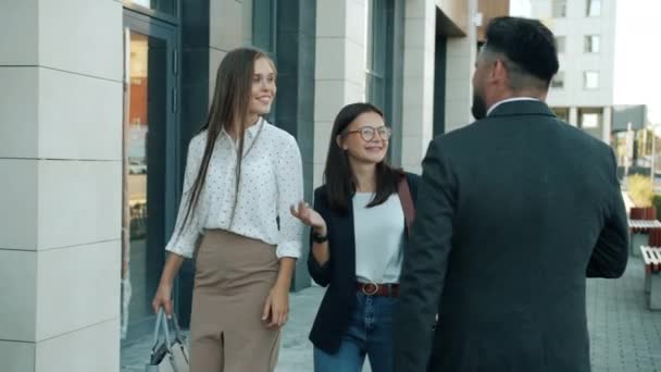 Movimiento lento de las señoritas alegres en ropa formal caminando al aire libre en las aceras saludando colega masculino — Vídeos de Stock