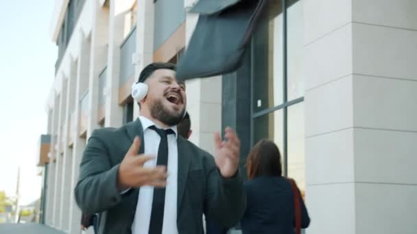 Movimiento lento del hombre de negocios despreocupado con auriculares caminando al aire libre en la ciudad disfrutando de la música — Vídeos de Stock