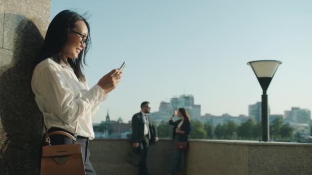 Powolny ruch pięknej bizneswoman azjatyckiej za pomocą smartfona stojącego na zewnątrz w pobliżu biura buiding w letni dzień — Wideo stockowe