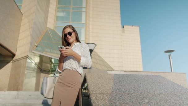 Atrakcyjna młoda kobieta korzystająca ze smartfona stojącego na zewnątrz w pobliżu biurowca, podczas gdy biznesmeni przechodzą obok — Wideo stockowe