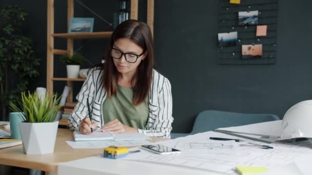 Young woman architect working with blueprint making notes then talking on mobile phone in office — Stock Video