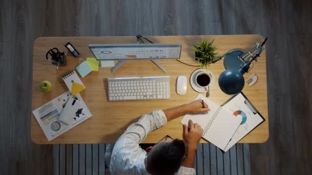 Vista de ángulo alto del hombre de negocios que trabaja en la mesa en la escritura de la oficina en el cuaderno mirando la pantalla del ordenador — Vídeos de Stock