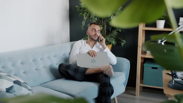 Φιλόδοξος νέος ελεύθερος επαγγελματίας που εργάζεται από το σπίτι συζητώντας τις επιχειρήσεις στο κινητό τηλέφωνο και χρησιμοποιώντας το lap-top — Αρχείο Βίντεο