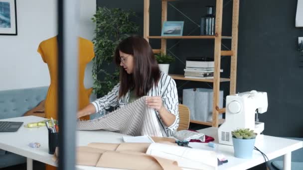 Młoda kobieta ubrania projektant pomiaru tkaniny i za pomocą laptopa pracy w mieszkaniu — Wideo stockowe