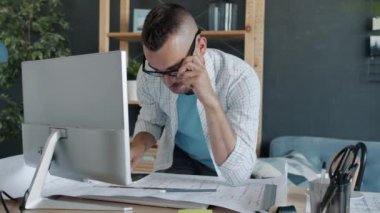 Genç mühendis cep telefonuyla iş tartışıyor sonra da bilgisayar ekranına bakarak plan üzerinde çalışıyor.