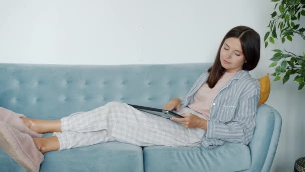 Втомилася дівчина в повсякденному одязі, використовуючи ноутбук, що працює, а потім завантажується на диван вдома — стокове відео