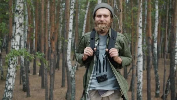 Porträt eines gut gelaunten Touristen, der in lässiger Kleidung mit Rucksack und Kamera im Wald steht — Stockvideo