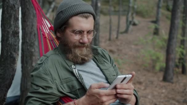 Młody człowiek turysta za pomocą smartfona dotykanie ekranu i relaks w hamaku w lesie — Wideo stockowe