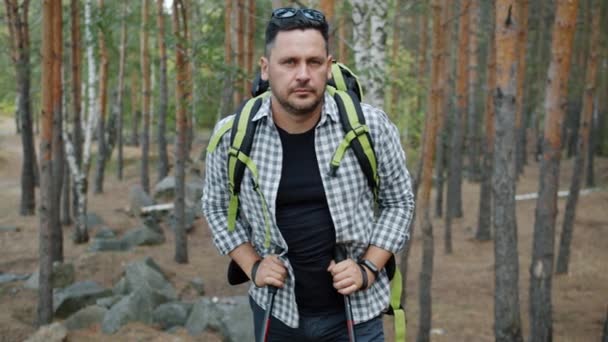 Zeitlupenporträt eines Mannes, der mit Rucksack und Wanderstöcken im Wald steht und mit ruhiger Miene in die Kamera blickt — Stockvideo