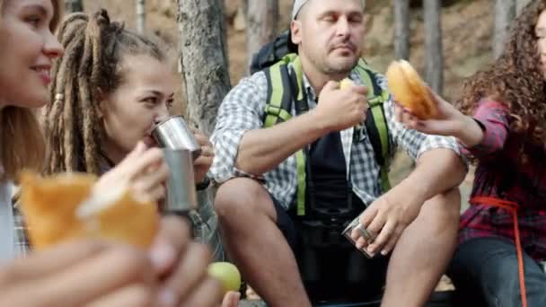 Grupa szczęśliwych turystów jedzących kanapki i pijących herbatę z metalowych kubków rozmawiających cieszących się wakacjami w lesie — Wideo stockowe