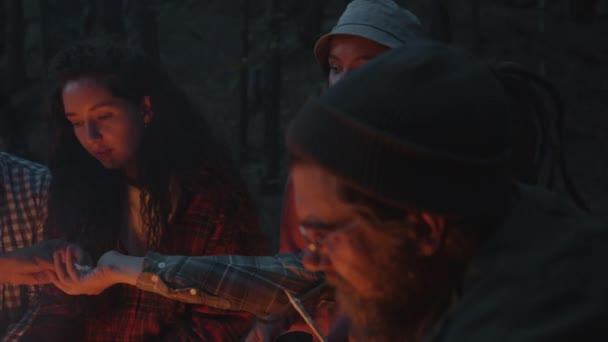 Vreugdevolle toeristen mannen en vrouwen roosteren marshmallow boven vuur praten lachen in donker bos — Stockvideo