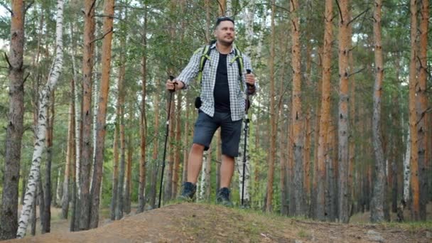 Porträt eines aktiven jungen Mannes, der mit Nordic-Walking-Stöcken und Rucksack im Wald steht und sich umschaut — Stockvideo