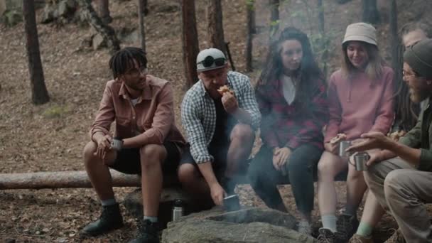 Portret van vrolijke jongeren die rond vuur eten in het bos en lachen met een hond die rondrent — Stockvideo