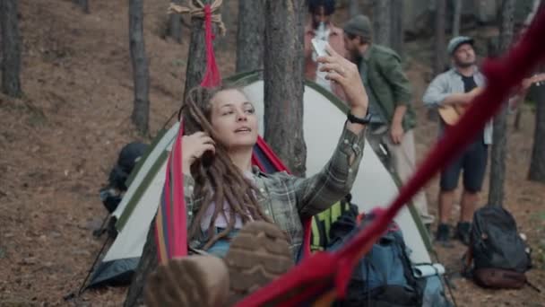 Привлекательная девушка турист делает селфи со смартфоном камеры отдыха в гамаке в кемпинге в лесу — стоковое видео