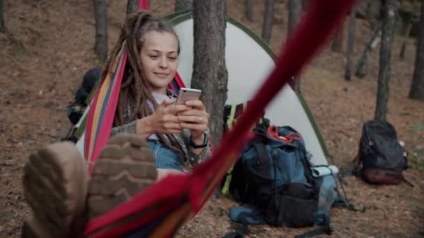 Vidám fiatal hölgy raszta pihentető függőágyban az erdőben, és okostelefon mosoly — Stock videók