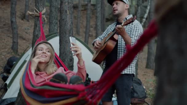 Gadis pirang mengambil foto selfie dengan kamera smartphone berayun di tempat tidur gantung di hutan sementara pacar bermain gitar — Stok Video
