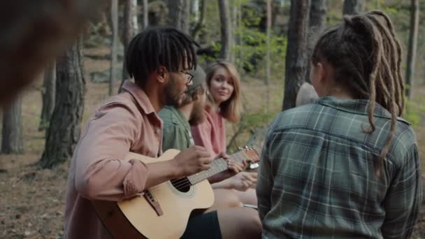 Powolny ruch wesołych przyjaciół wielonarodowa grupa gra na gitarze i śpiewa piosenki na świeżym powietrzu w lesie — Wideo stockowe