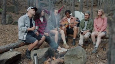 Ormanda kamp ateşinin yanında gitar çalıp şarkı söyleyen genç turistlerin yavaş çekimde portresi.