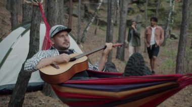 Yürüyüşçüler arka planda yürürken hamakta gitar çalan neşeli adam.