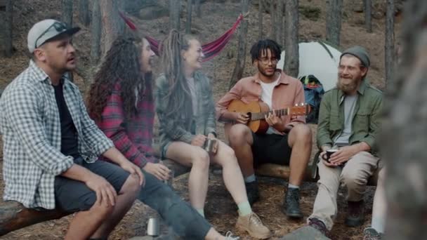 Весёлые друзья-студенты поют, играя на гитаре, смеясь, сидя у костра в лесу — стоковое видео