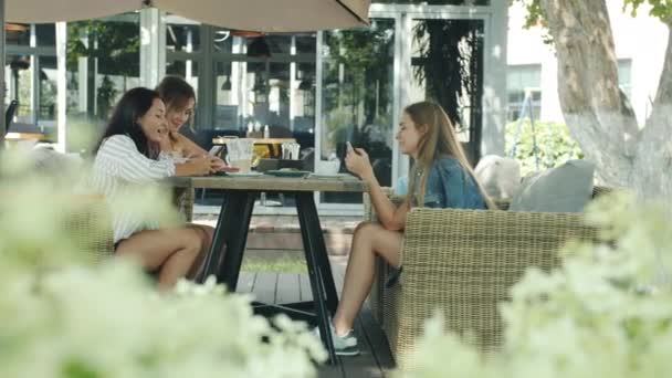 Side view of young ladies friends talking and using smartphones sitting at table in outdoor cafe — Stock Video