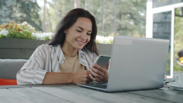Veselý mladý businesswoman pomocí smartphone smích sedí u stolu v venkovní kavárně — Stock video