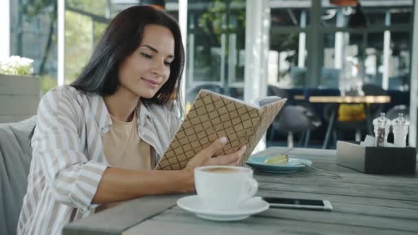 Portret van een mooie jonge brunette leesboek genieten van literatuur in cafe op zomerdag — Stockvideo