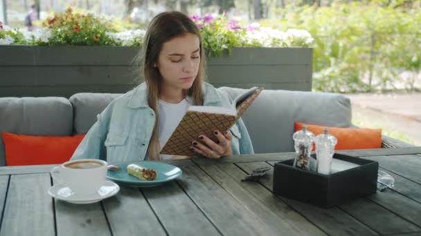 Retrato de la hermosa rubia joven disfrutando de libro sentado en la mesa en la cafetería al aire libre — Vídeos de Stock