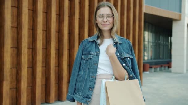 Vrolijke jonge dame shopaholic buiten staan met papieren tassen dragen trendy kleding — Stockvideo