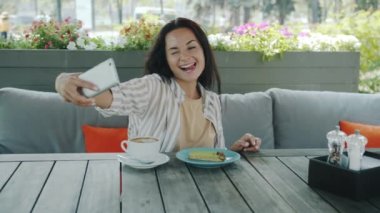 Akıllı telefon kamerasına poz veren neşeli genç bayanın açık kafede selfie çektiği yavaş çekim portresi.