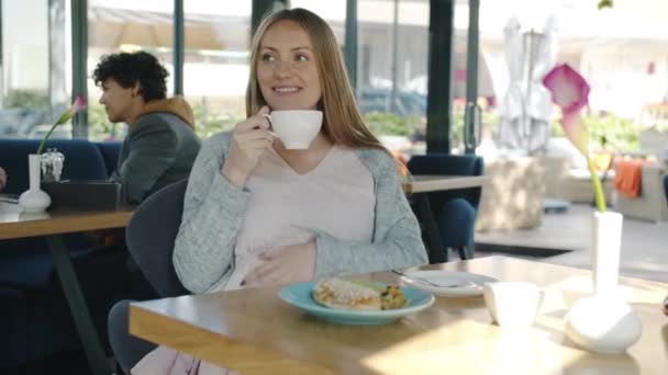 Веселая беременная женщина наслаждается едой в кафе, сидя за столом с едой и напитками, улыбаясь в летний день — стоковое видео
