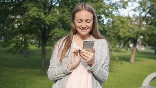 未来的母亲用智能手机独自站在城市公园里，微笑着享受现代设备和大自然 — 图库视频影像