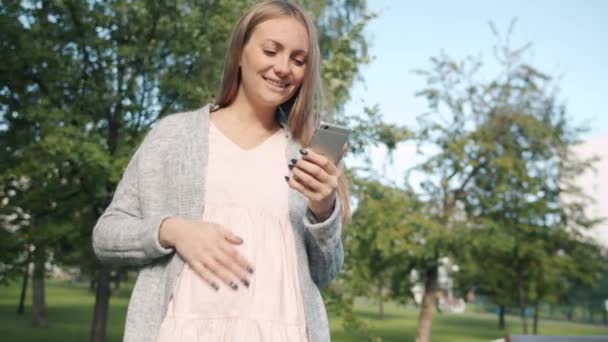 Mãe esperando alegre olhando para a tela do telefone inteligente e sorrindo de pé no parque da cidade no verão — Vídeo de Stock