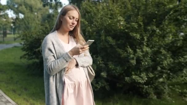 Joven embarazada rubia caminando en el parque de la ciudad grabación de mensajes de audio con teléfono inteligente — Vídeo de stock