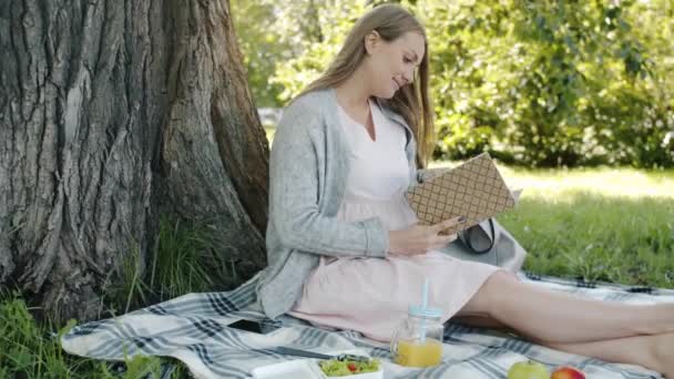 Hermosa joven futura madre leyendo libro sentado en cuadros durante el picnic en el parque en el día de verano — Vídeo de stock