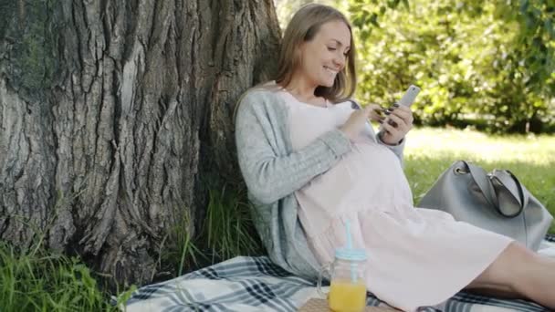 Bella mujer embarazada disfrutando de la aplicación de teléfono inteligente y beber jugo relajante en el césped en el parque de la ciudad — Vídeo de stock