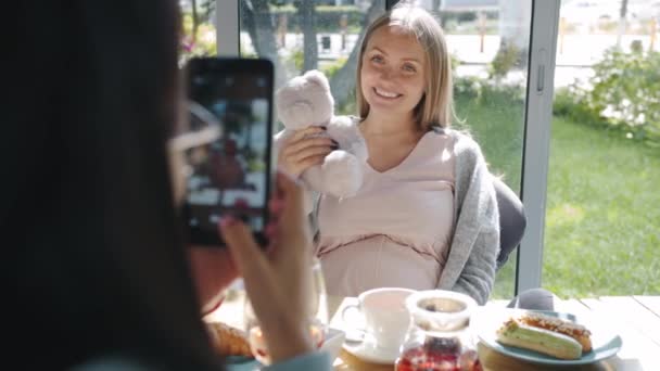 お友達とカフェで写真を撮っている間にテディベアとスマートフォンカメラのためにポーズをとって幸せな妊婦 — ストック動画