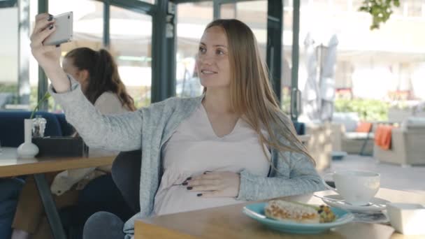 Радостная беременная женщина делает селфи, развлекаясь в современном летнем кафе, сидя за столом одна. — стоковое видео
