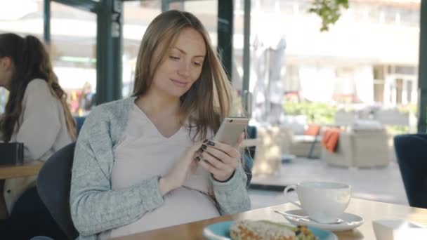Веселая беременная блондинка с помощью смартфонов, сидящая за столом в современном кафе — стоковое видео