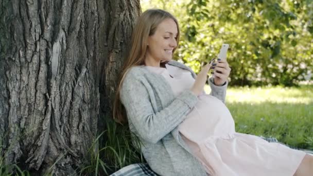 Retrato de futura madre alegre usando teléfono inteligente sentado bajo el árbol en el parque de la ciudad — Vídeo de stock